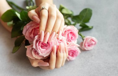Pembe manikürlü bir kadının elleri pembe güller tutuyor ve elmas yüzüğünü gösteriyor.