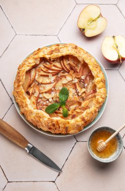 Badem gevreğiyle süslenmiş taze pişmiş elma galeti, yanında bal ve elma dilimleri, şık bir fayans masasında dinleniyor, tadını çıkarmaya hazır.