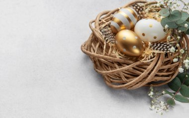 Paskalya yumurtaları gri bir arka planda kuş tüyü ve çiçeklerle süslenmiş dekoratif bir yuvada yatar.