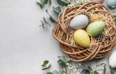 Hasır bir yuvada, çiçekler ve okaliptüs yapraklarıyla çevrili saman ve tüylerle boyanmış paskalya yumurtaları.