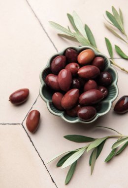 Kalamata zeytinleri ve zeytin dalları yeşil bir kâsede fayanslı bir yüzeyde sergilenerek Akdeniz mutfağı sahnesi oluşturulur.