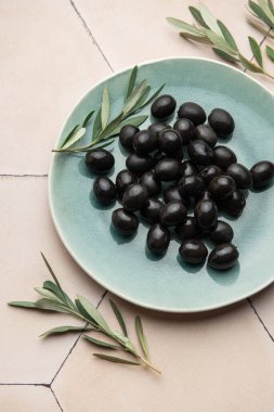 Siyah zeytinler seramik bir tabakta seramik bir yüzeyde zeytin dallarıyla servis edilir ve Akdeniz mutfağı sahnesi oluşturulur.