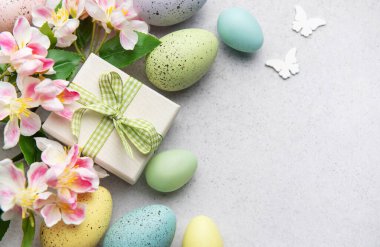 Renkli paskalya yumurtaları, yeşil kurdeleli hediye kutusu ve bahar çiçekleri. Fotokopi alanı olan gri arka planda neşeli bir sınır oluşturuyor.