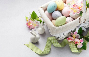 Renkli paskalya yumurtaları ve bahar çiçekleri açık arkaplanda beyaz bir hasır sepetten taşar.