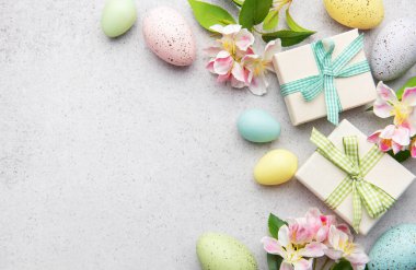 Renkli paskalya yumurtaları ve hediye kutuları bahar çiçekleriyle süslenmiş gri bir arka planda yatıyor.