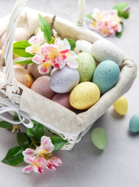 Renkli paskalya yumurtaları ve bahar çiçekleri açık arkaplanda beyaz bir hasır sepetten taşar.