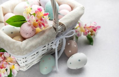 Renkli paskalya yumurtaları ve bahar çiçekleri açık arkaplanda beyaz bir hasır sepetten taşar.