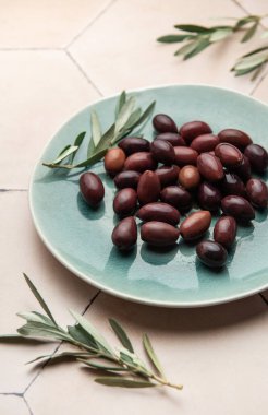 Taze zeytin dallarıyla süslenmiş Kalamata zeytinleri, modern bir tabakta fayanslı bir yüzey üzerinde servis edilerek, şık ve iştah açıcı bir sunum oluşturur.