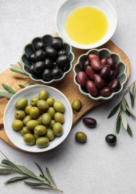 Kase dolusu yeşil, siyah ve kalamata zeytinyağı ve zeytin dalları, ahşap bir kesme tahtasının üzerinde duruyor.