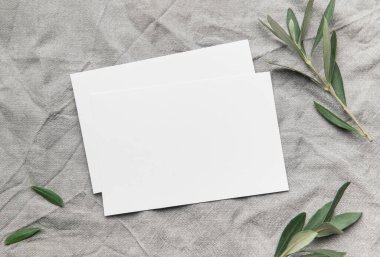 Taze zeytin dallarıyla süslenmiş gri keten kumaş üzerine iki boş kart yerleştiriliyor. Huzurlu ve minimalist bir sahne yaratıyor.