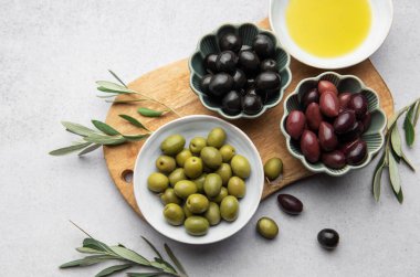 Kase dolusu yeşil, siyah ve kalamata zeytinyağı ve zeytin dalları, ahşap bir kesme tahtasının üzerinde duruyor.