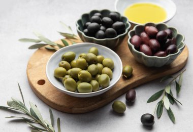 Kase dolusu yeşil, siyah ve kalamata zeytinyağı ve zeytin dalları, ahşap bir kesme tahtasının üzerinde duruyor.