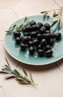 Siyah zeytinler seramik bir tabakta seramik bir yüzeyde zeytin dallarıyla servis edilir ve Akdeniz mutfağı sahnesi oluşturulur.