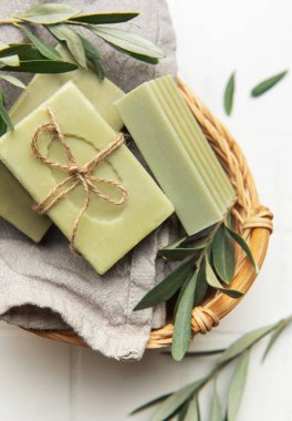 Beyaz fayanslı sepetin içinde zeytin dalı olan ev yapımı zeytin sabunları doğal hijyeni destekliyor.