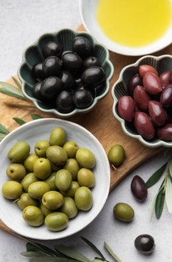 Kase dolusu yeşil, siyah ve kalamata zeytinyağı ve zeytin dalları, ahşap bir kesme tahtasının üzerinde duruyor.