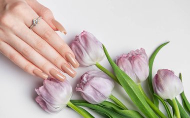 Bej renkli manikürlü ve elmas yüzüklü bir kadın. Mor lale buketinin yanında cilalanmış tırnakları var.