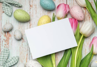 Renkli paskalya yumurtaları, laleler ve tavşan kulakları kırsal ahşap bir arka planda boş bir kartı sarıp neşeli bir bahar ayarı yaratır.