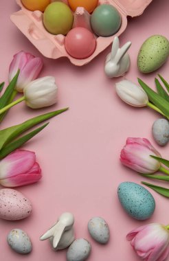 Paskalya yumurtaları, seramik tavşanlar ve laleler pembe arka planda fotokopi alanı olan bir çerçeve oluşturuyorlar.