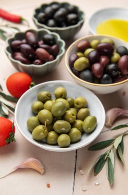 Kâselerde çeşitli zeytinler sunulurken, domates, sarımsak, zeytinyağı, tuz ve biber gibi taze malzemeler davetkar bir aşçılık sahnesi yaratıyor.