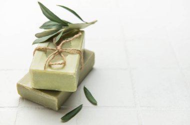 Two bars of olive oil soap, tied with twine and decorated with olive branches, resting on white tiles, promoting natural body care