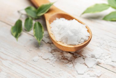 Tahta kaşıkta duran magnezyum klorür tanecikleri ve beyaz ahşap masada yeşil yapraklar sağlığını ve sağlığını destekliyor.