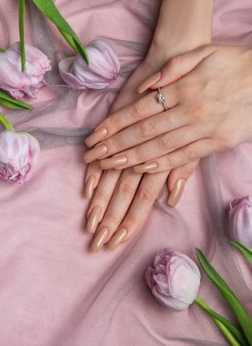 Çıplak manikür ve pırlanta yüzüklü kadın elleri pembe kumaşın üzerinde lalelerin yanında yatıyor.