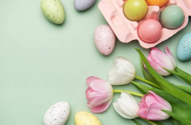 Renkli paskalya yumurtaları ve pembe ve beyaz laleler pastel yeşil arka planda fotokopi alanı ile bahar hissi yaratıyor.