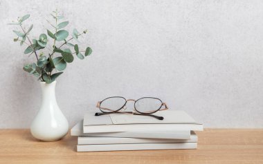 Gözlük, bir vazo okaliptüs dalının yanındaki kitap yığınının üzerinde, huzurlu bir okuma atmosferini somutlaştırıyor.