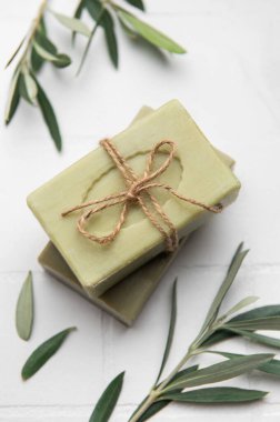 İki çubuk zeytin sabunu sicimle bağlanmış, zeytin dallarıyla beyaz bir fayans üzerinde dinleniyor, doğal vücut bakımını ve hijyeni teşvik ediyor.