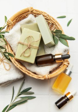 El yapımı zeytin sabunları, gerekli yağ şişeleri, ve beyaz fayans zemininde hasır bir sepete yerleştirilmiş taze zeytin dalları, sakin bir spa ortamını çağrıştırıyor.