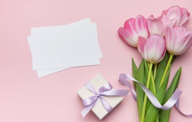 Pembe laleler, leylak kurdeleyle bağlanmış bir hediye kutusu ve pastel pembe arka planda boş kartlar içeren narin düz bir kompozisyon.