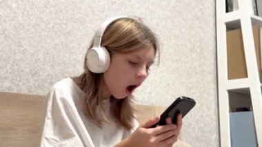 Akıllı telefon ve kulaklık kullanan, yatakta oturan, farklı duygular gösteren şaşırmış bir kız.