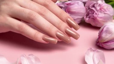 Pembe arka planda mor lalelerin yanında bej renkli tırnaklarını gösteren bir kadın.