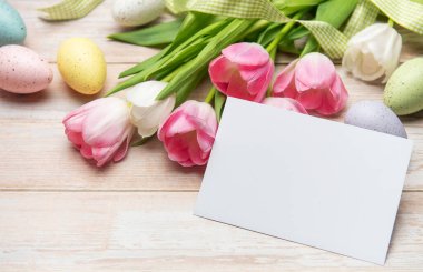 Paskalya yumurtaları ve laleler, mesajınız için boş bir tebrik kartıyla ahşap bir masada yatıyor.