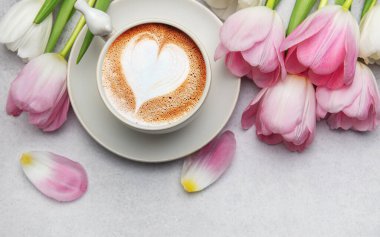 Bir fincan kapuçino ile latte sanatı ve pembe laleler gri masada uzanmış, aşkı ve baharı kutluyorlar.