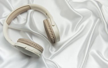 Bej ve beyaz kablosuz kulaklıklar lüks beyaz ipek arka planda dinleniyor, zarafet hissi yaratıyor ve yüksek kaliteli ses deneyimi