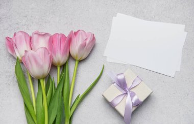 Pembe laleler, leylak kurdeleli hediye kutusu ve boş tebrik kartları gri desenli arka planda duruyor, neşeli bir hava yaratıyor.