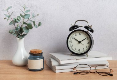 Bir çalar saat, kitaplar, gözlükler, bir mum ve bir vazoda okaliptüs dalları içeren minimalist ofis masası kurulumu