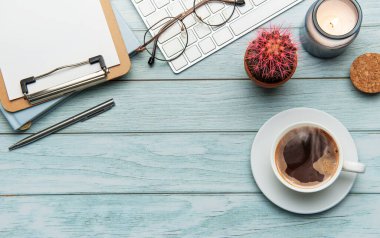 Açık mavi ahşap zemin üzerinde kahve, klavye, not defteri, bardak, kalem, mum ve kaktüs ile ofis masasının düz kompozisyonu.