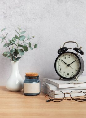 Bir çalar saat, kitaplar, gözlükler, bir mum ve bir vazoda okaliptüs dalları içeren minimalist ofis masası kurulumu
