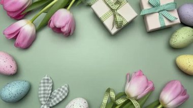 Yeşil arka planda pembe laleler, hediye kutuları ve pastel renkli yumurtaların yer aldığı Paskalya düzlemi, bayram sahnesi yaratıyor.