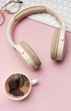 Kablosuz kulaklıklar, klavye ve gözlükler pembe arka planda bir fincan kahvenin yanında, ev dekorasyonundan rahatlatıcı bir çalışmayı temsil ediyor.