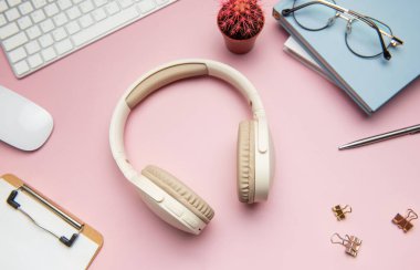 Kablosuz kulaklıklar, klavye, fare, kırtasiye, gözlük ve pembe arka planda bir kaktüs içeren modern çalışma alanı