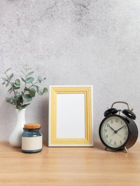 Boş çerçeveli modern ev dekoru, çalar saat, okaliptüs dalları ve gri bir duvara karşı tahta bir masanın üzerinde boş etiketli kavanoz.