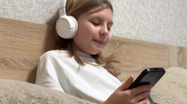 Kulaklık takan kız yatakta dinlenirken akıllı telefonuyla müzik dinlemekten hoşlanır.