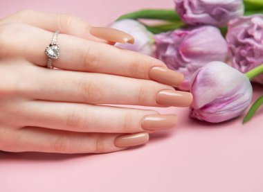 Kadın eli, kusursuz çıplak manikür ve pembe arka planda mor lalelerin yanında kalp şeklinde elmas yüzük sunar.