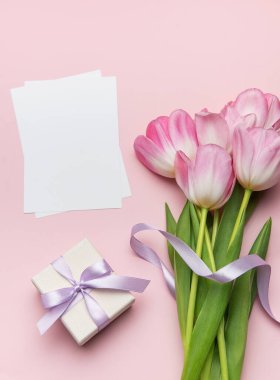 Pembe laleler, leylak kurdeleyle bağlanmış bir hediye kutusu ve pastel pembe arka planda boş kartlar içeren narin düz bir kompozisyon.