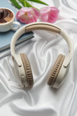 Kablosuz kulaklıklar, bir fincan kahve, bir kitap ve pembe laleler beyaz ipek üzerinde rahatlatıcı bir atmosfer yaratır.