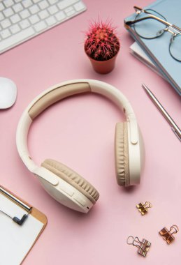 Kablosuz kulaklıklar, klavye, fare, kırtasiye, gözlük ve pembe arka planda bir kaktüs içeren modern çalışma alanı