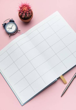 Kalem, alarm saati ve kaktüsle birlikte planlama ve üretkenliği temsil eden pembe bir arkaplan üzerine açık bir defter.